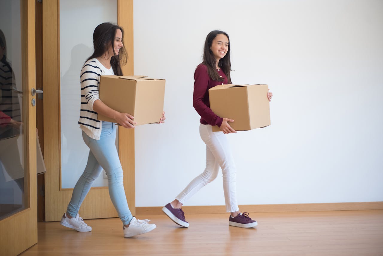Two young women carrying cardboard boxes indoors, illustrating teamwork and moving in.