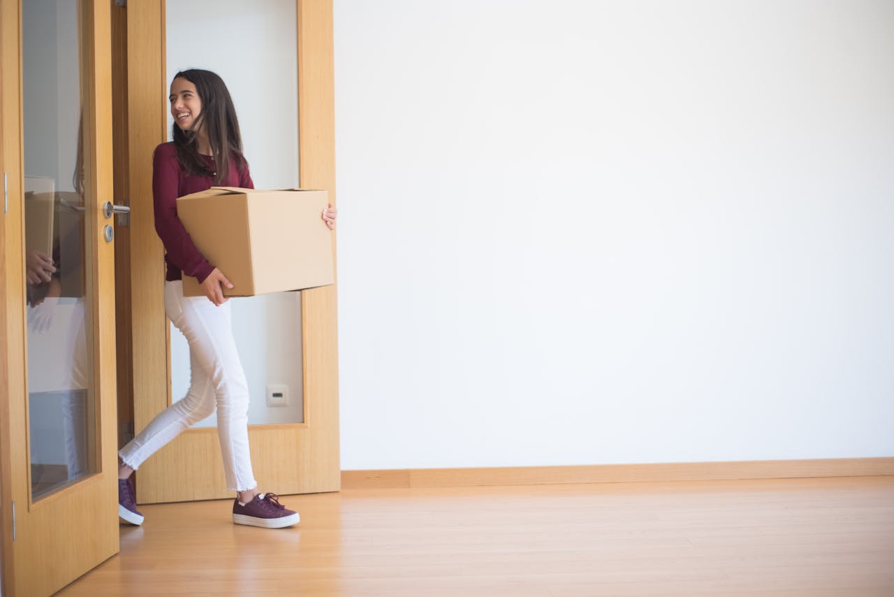 A woman enters a new apartment carrying a cardboard box, symbolizing moving in.
